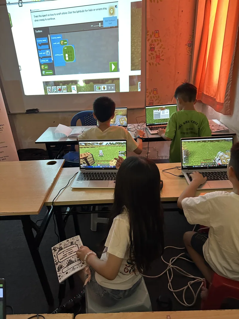 Students working on Minecraft projects with a coding demonstration projected at the front of the classroom